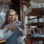 Idosa recebe orientação de uma farmacêutica diante das prateleiras de uma farmácia.