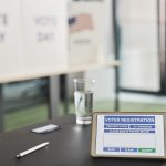 Tablet com tela de cadastro eleitoral em uma seção de votação interna, ao lado de placas que indicam o dia da votação.