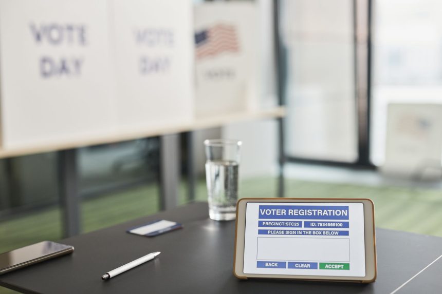 Tablet com tela de cadastro eleitoral em uma seção de votação interna, ao lado de placas que indicam o dia da votação.