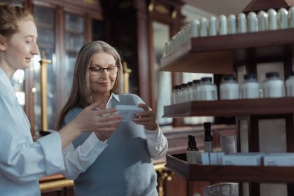 Idosa recebe orientação de uma farmacêutica diante das prateleiras de uma farmácia.