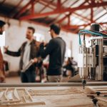 Três homens conversam em uma oficina de marcenaria, com uma máquina CNC ao fundo.