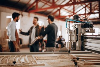 Três homens conversam em uma oficina de marcenaria, com uma máquina CNC ao fundo.