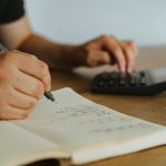Mãos fazem cálculos em um caderno ao lado de uma calculadora, representando planejamento financeiro.