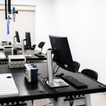 Laboratório de eletrônica com computadores, monitores e equipamentos técnicos sobre bancadas.