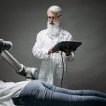 Cientista monitora um braço robótico enquanto ele realiza um procedimento em uma pessoa deitada em um ambiente de pesquisa.