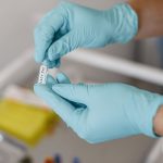 Mãos enluvadas seguram um frasco de vacina em ambiente clínico