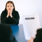 Mulher participa de entrevista de emprego diante de dois recrutadores, com o currículo sobre a mesa.