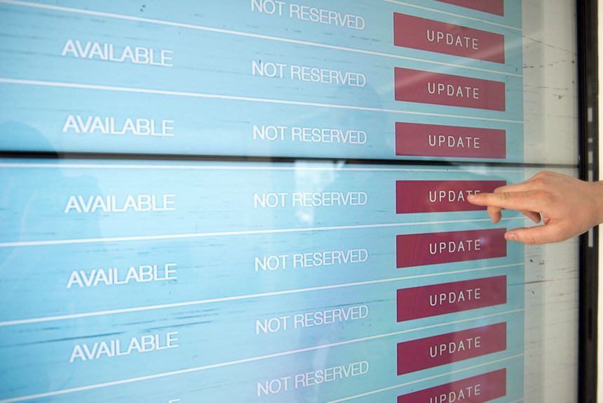Mão interage com uma tela sensível ao toque que exibe opções digitais de disponibilidade e atualização de informações.